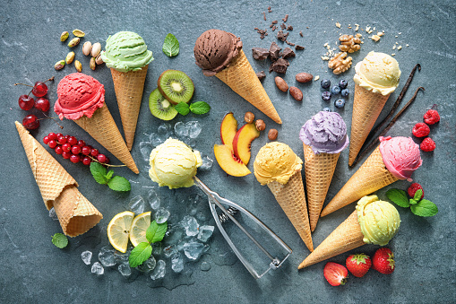 Various varieties of ice cream in cones with mint, blueberry, strawberry,  pistachio, cherry, chocolate and scoop on gray stone background