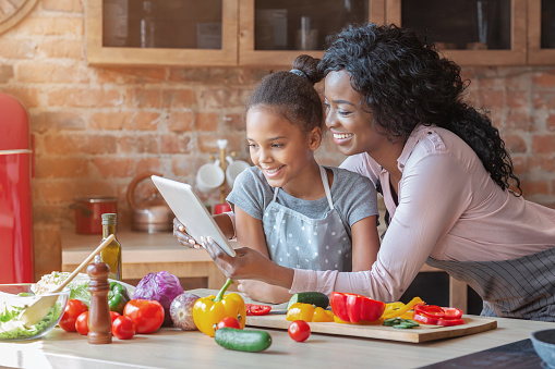 Happy parenthood. Black woman and her cute daughter reading recipe on tablet, cooking at kitchen, copy space