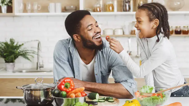 African girl feeding dad in kitchen, giving him cherry tomato while cooking salad, copy space