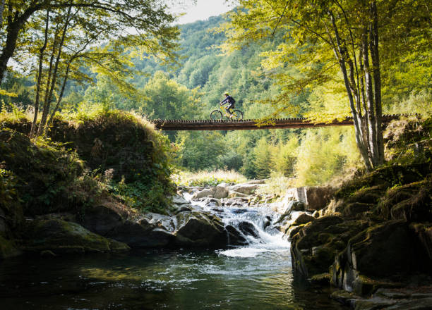 Woman cycling cross country in nature, side view of woman biker on wooden bridge over the waterfall in beautiful mountain location.