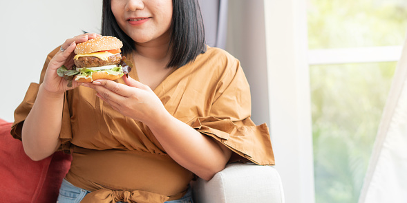 Hungry overweight woman smiling and holding hamburger and sitting in the living room, her very happy and enjoy to eat fast food. Concept of binge eating disorder (BED).