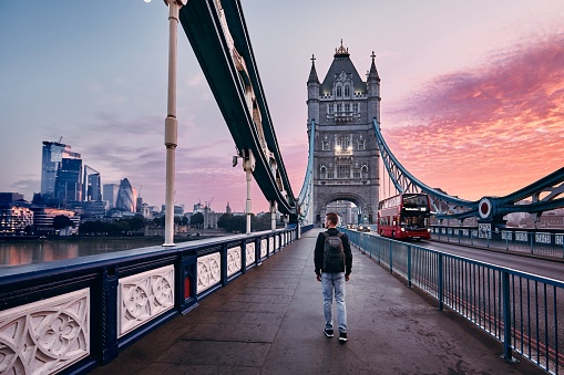 Young man with backpack walking on Tower Bridge against cityscape with skyscrapes at colorful sunrise. London, United Kingdom