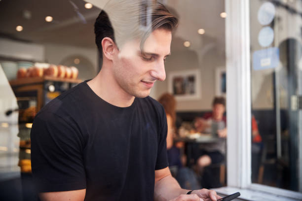 istock Young Man In Coffee Shop Looking At Mobile Phone Viewed Through Window 1173600628