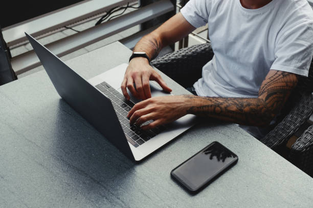 istock Handsome guy with tattooed hands dressed white shirt working on laptop, male typing text on notebook keyboard. Freelancer working on new startup project in city cafe with free wireless zone 1173614011