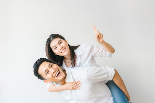 Happy Asian couple in love having fun piggyback ride and point at empty copyspace on white wall background
