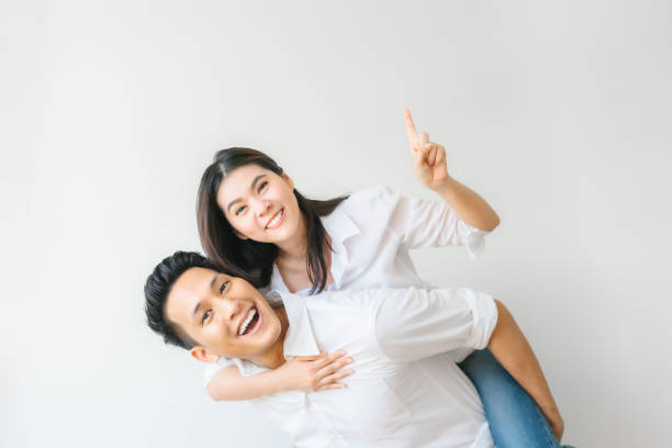 couple in love having fun piggyback ride and point at empty copyspace Happy Asian couple in love having fun piggyback ride and point at empty copyspace on white wall background couple isolated wife husband stock pictures, royalty-free photos & images