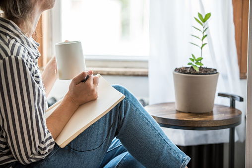 A young woman takes a break to do something analog like writing in her journal and drinking tea. This is a healthy practice for those who experience anxiety.