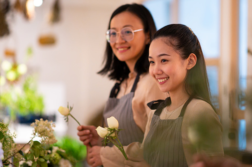 Florists examining white roses while arranging decoration. Females are wearing aprons while working together in workshop. Focus is on smiling woman during floral training class.