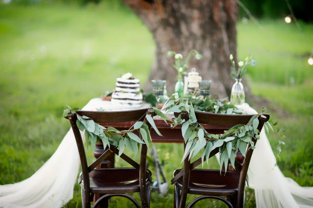istock Wedding decorated table for two with a white tablecloth decorated with floral composition and a cake on a background of grass and pine tree. Rustic style. 1173623305
