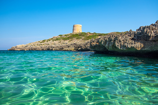 View on the beach Calo Roig with crystal water and the Defense tower Torre de Defensa de Alcaufar on Menorca, Balearic Islands, Spain.