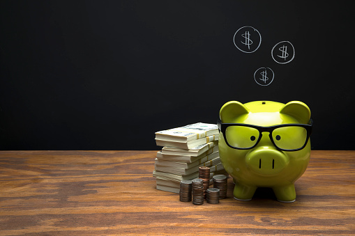 This is a photograph of a green Piggy Bank sitting on a desk a with a chalk drawing of a coin shape symbol sign on Black Chalkboard Background. This image could relate to savings and retirement.