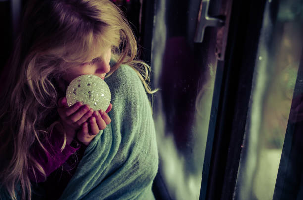 lovely blond girl sitting by the window with christmas bauble and looking from the window