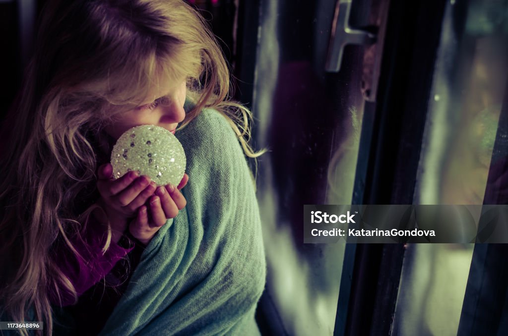 child with christmas bauble looking through window lovely blond girl sitting by the window with christmas bauble and looking from the window Sadness Stock Photo