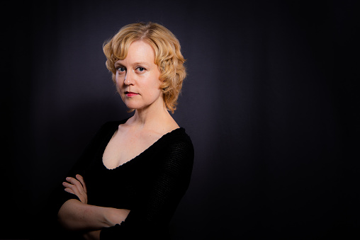 Portrait of a confident woman in her forties with short blond hair, in front of a dark background.