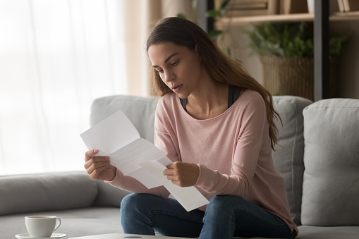 Concentrated girl sit on couch holding paper document in hands reading carefully examining, focused millennial woman study received letter, check post paperwork correspondence in living room