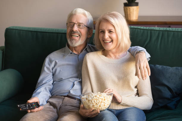 istock Happy senior couple holding remote control snack laughing watching tv 1173666976