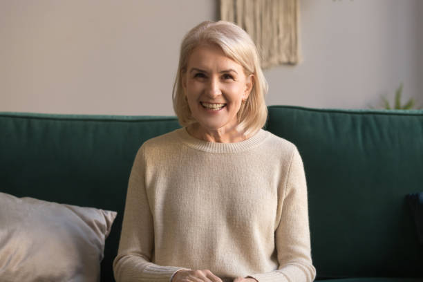 smiling middle aged woman talking to camera sit on couch - senior adult happiness computer looking at camera imagens e fotografias de stock