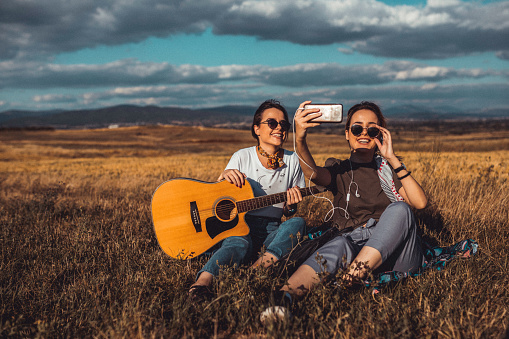 Two teenage girls playing guitar and photographing with a smart phone during a picnic on a sunny summer day