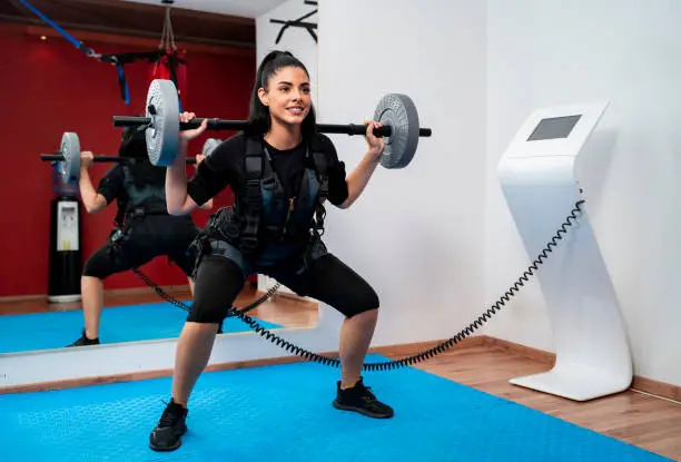 Female doing squats exercise with weights attached to EMS machine and smiling