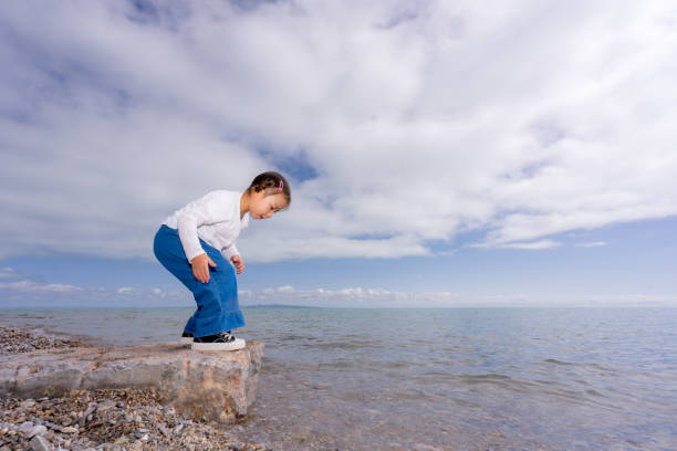 A young child walks barefoot along a sandy beach, wearing a striped dress, as the sun sets in the background. The peaceful setting creates a serene atmosphere for exploration.