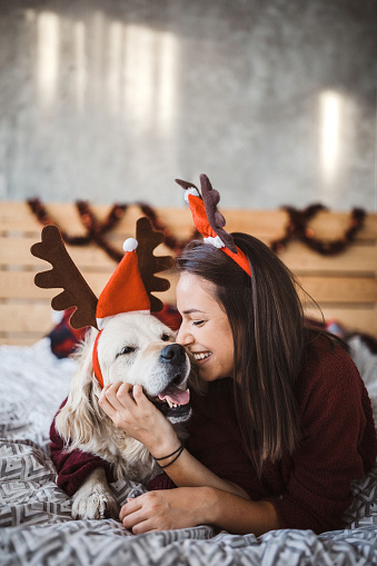 Cheerful girl lying on bed with her dog, both wearing reindeer  antlers, cuddling