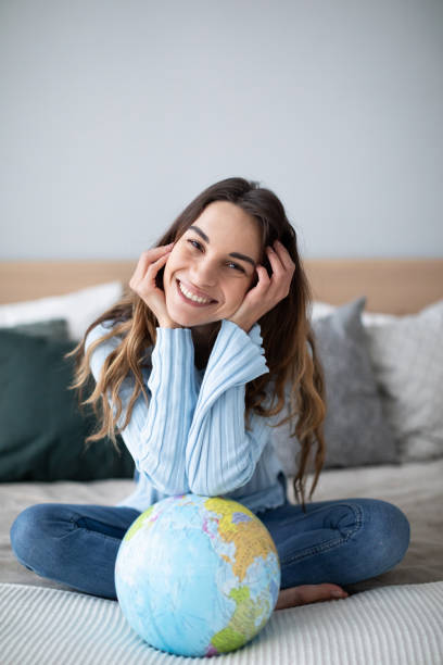 ベッドの上に座ってカメラを見ている地球儀を持つ幸せな笑顔の女性。 - travel globe student journey ストックフォトと画像