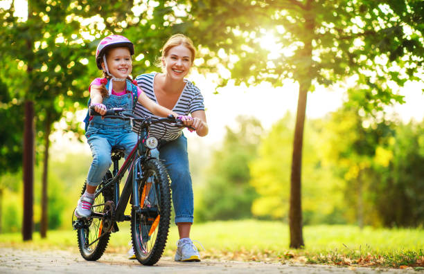 madre feliz familia enseña a su hija a montar en bicicleta en el parque - casco de deportes fotos fotografías e imágenes de stock