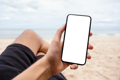 Mockup image of a hand holding and showing black mobile phone with blank desktop screen while sitting on the beach chair