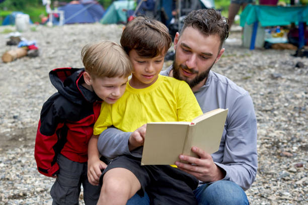 A man reads a book to two boys while camping. The eldest son sits on his father