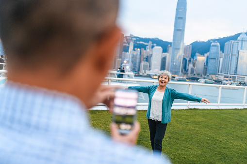 Over the shoulder view of mature Chinese man taking cheerful senior woman selfie with Hong Kong cityscape in background.