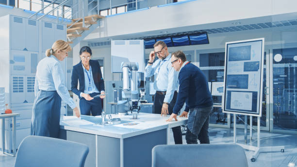 istock Diverse Team of Industrial Robotics Engineers Gathered Around Table With Robot Arm, They Manipulate and Program it to Pick Up and Move Metal Component. Bright Facility, Creative People 1173740415