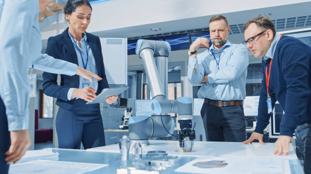 istock Diverse Team of Industrial Robotics Engineers Gathered Around Table With Robot Arm, They Manipulate and Program it to Pick Up and Move Metal Component. Bright Facility, Creative People 1173740424