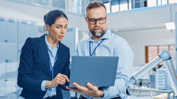 istock Female Chief Engineer Talks with Male Electronics Specialist, Explaining Things, He Holds on Laptop Computer. Modern and Bright Office with Stylish People and Working Robot Arm in Background 1173740546
