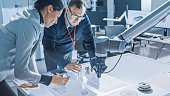 Stylish Female Engineer and Male Specialist Talking Lean on the Table Work with Blueprints, Documents and Tablet Computer. Working on Program and Manipulation of Robot Arm Movements. Modern Facility