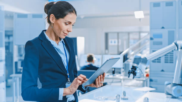 istock Stylish Female Industrial Robotics Engineer Uses Tablet Computer to Program and Manipulate Robot Arm for Lifting, Moving Objects. Technology Research Facility for Machine Learning and Cloud Computing 1173740579