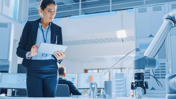 istock Stylish Female Industrial Robotics Engineer Uses Tablet Computer to Program and Manipulate Robot Arm for Lifting, Moving Objects. Technology Research Facility for Machine Learning and Cloud Computing 1173740581