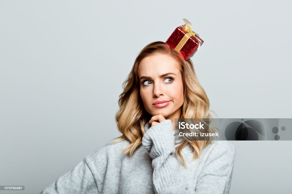 Christmas portrait of cute beautiful woman, close up of face stock photo Mid adult beautiful woman wearing sweater standing against grey background, looking up with hand on chin. Gift Stock Photo