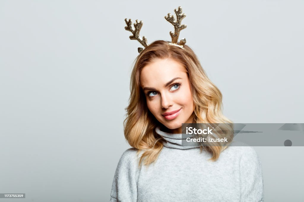 Christmas portrait of cute beautiful woman, close up of face stock photo Mid adult beautiful woman wearing sweater and reindeer horns standing against grey background, looking up. Christmas Stock Photo
