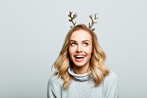 Mid adult beautiful woman wearing sweater and reindeer horns standing against grey background, looking up and laughing.