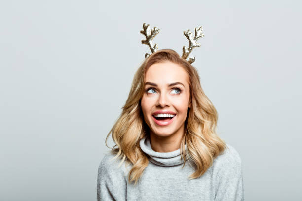 Christmas portrait of excited beautiful woman, close up of face stock photo Mid adult beautiful woman wearing sweater and reindeer horns standing against grey background, looking up and laughing. christmas excitement stock pictures, royalty-free photos & images