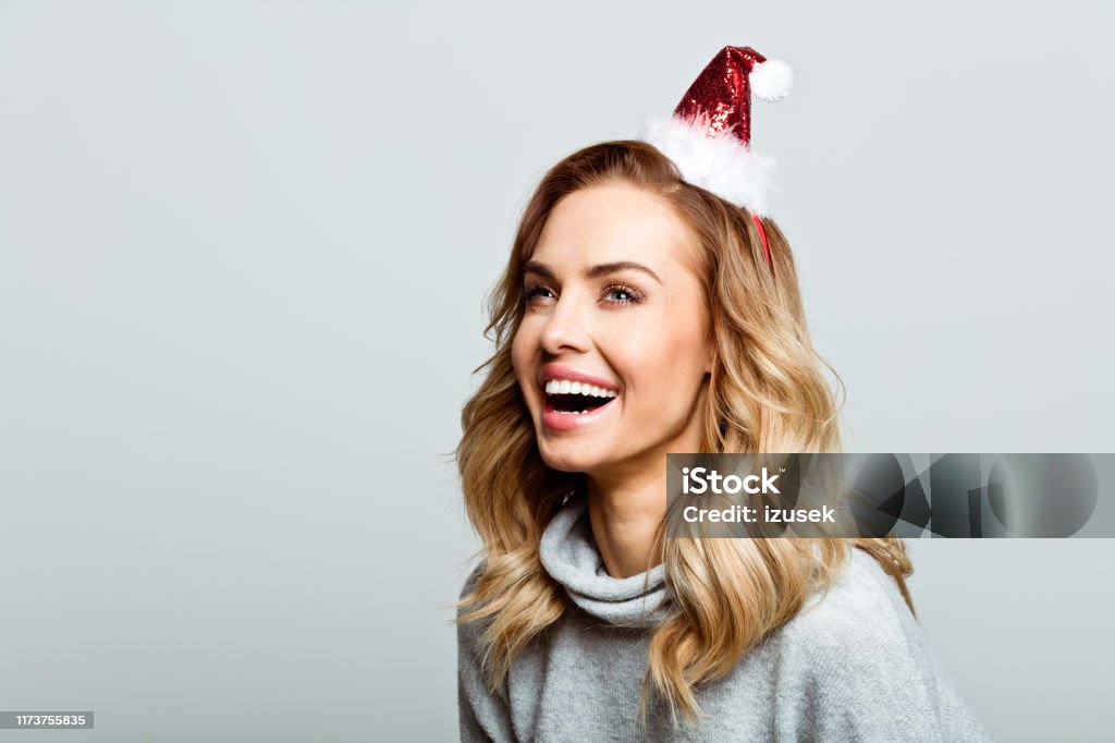 Christmas portrait of excited beautiful woman, close up of face stock photo Mid adult beautiful woman wearing sweater and Santa Claus hat standing against grey background, looking up and laughing. Christmas Stock Photo