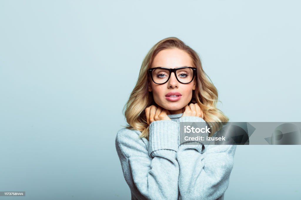 Winter portrait of beautiful blonde woman, close up of face stock photo Mid adult beautiful woman wearing sweater and glasses standing against grey background, looking at camera. Eyeglasses Stock Photo