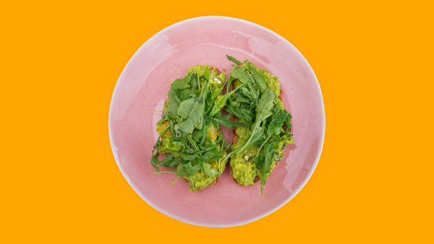 istock Two slices of mashed avocado toast garnished with fresh rocket or arugula salad set on a pink plate against a bright orange background 1173759351