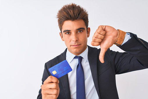Young handsome businessman wearing suit holding credit card over isolated white background with angry face, negative sign showing dislike with thumbs down, rejection concept