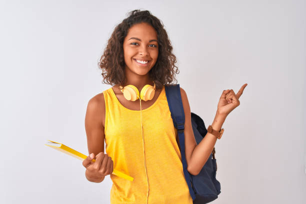 mujer estudiante brasileña con mochila sosteniendo cuaderno sobre fondo blanco aislado muy feliz señalando con la mano y el dedo hacia un lado - estudiante fotos fotografías e imágenes de stock