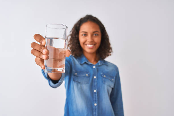 junge brasilianische frau hält glas wasser stehend über isolierten weißen hintergrund mit einem glücklichen gesicht stehen und lächelnd mit einem selbstbewussten lächeln zeigt zähne - toothy smile relaxation white healthcare and medicine stock-fotos und bilder