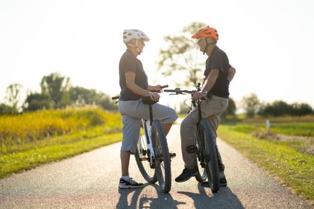 side view happy smiling senior couple on electric bicycles looking at each other on sunny summer day on cycle path through nature reserve
