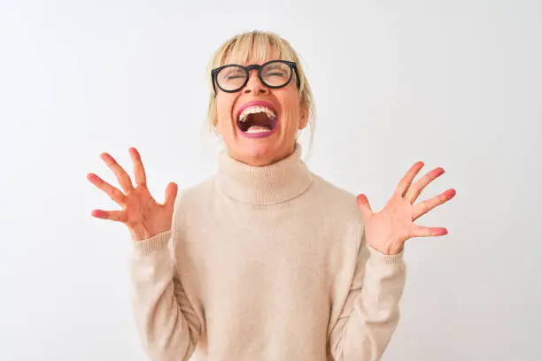 Photo of Middle age woman wearing turtleneck sweater and glasses over isolated white background celebrating mad and crazy for success with arms raised and closed eyes screaming excited. Winner concept