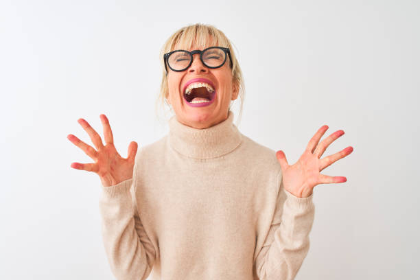 mulher da idade média que desgasta a camisola e os vidros do gola sobre o fundo branco isolado que comemora louco e louco para o sucesso com os braços levantados e os olhos fechados que gritam excited. conceito do vencedor - eufórico - fotografias e filmes do acervo