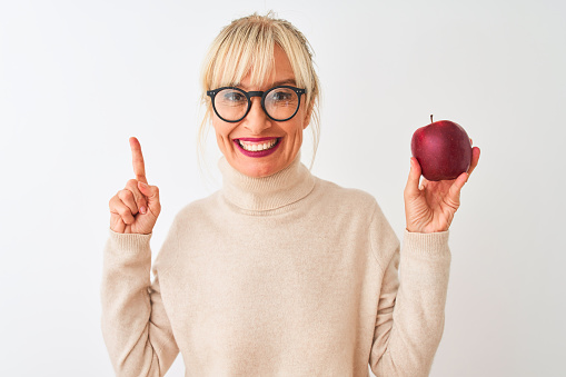 Middle age woman wearing glasses holding apple standing over isolated white background surprised with an idea or question pointing finger with happy face, number one